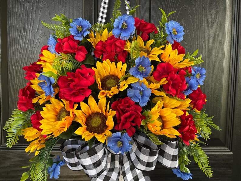 以下が含まれることがあります： ヒマワリ、赤いバラ、青い花が特徴の鮮やかな花のリース。緑の葉を含み、白黒のチェック柄のリボンでアクセントを加えています。ドアの装飾に最適です。