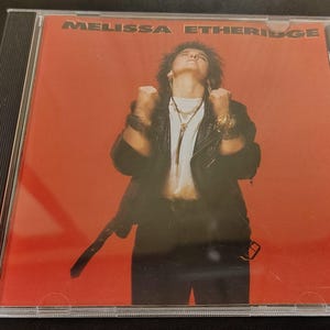 Peut inclure: Un CD de musique dans un boîtier en plastique transparent avec une pochette sur fond rouge et le texte MELISSA ETHERIDGE. L'image montre une personne aux cheveux courts foncés portant une veste en cuir noir, un haut blanc et des colliers.