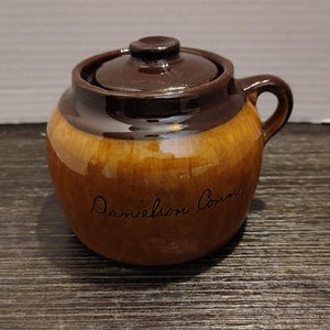 Può includere: Un pentolino rotondo in ceramica con smalto marrone lucido, dotato di coperchio coordinato con pomello e manico laterale. Il corpo del pentolino presenta la scritta nera manoscritta Danielson Court sulla parte anteriore.