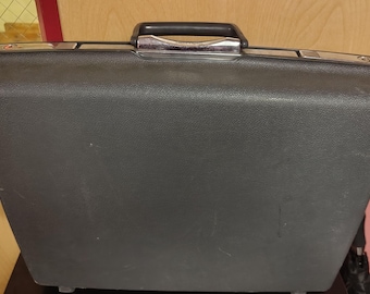 May include: A rectangular black hard-shell briefcase with a textured surface. The case features a black plastic handle with a central chrome-colored metal accent, and two silver-toned metal latches positioned on the top edge.