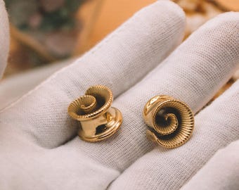 Puede incluir: Un par de dilatadores de oreja dorados con un diseño en espiral acanalado en la cara frontal. Las piezas de joyería son circulares con un acabado pulido y liso en los bordes exteriores y un patrón de remolino texturizado hacia el centro.