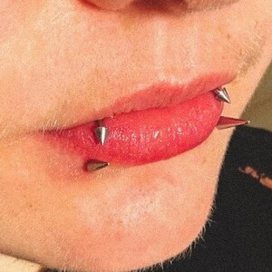 Puede incluir: Un conjunto de tres piercings labiales de metal plateado con extremos cónicos afilados. Las piezas se sitúan a lo largo del labio inferior, con dos puntas orientadas hacia afuera y una punta más pequeña centrada bajo la línea del labio.