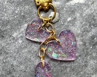 May include: A gold-toned metal keychain featuring two translucent purple heart-shaped charms and one small oval charm, all embedded with multicolored glitter and iridescent flakes, attached to a gold lobster clasp and a small flower charm.