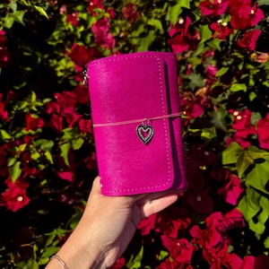 May include: A vibrant hot pink leather notebook cover with a textured hair-on-hide finish. The cover is secured by a thin pink elastic band featuring a small, silver-toned heart-shaped charm with embedded clear crystals centered on the front.