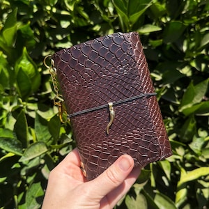 May include: A dark brown leather notebook cover featuring a textured snakeskin pattern. The cover is secured with a thin black elastic band adorned with a small gold-toned horn-shaped charm. A gold-toned metal chain with a clasp is attached to the side.