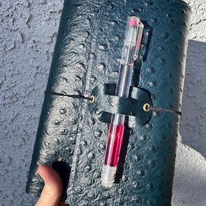 May include: A dark teal leather journal with a textured surface and a black elastic closure. A clear pen with pink ink is inserted into a leather pen holder on the journal. The journal is held in a hand.