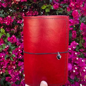 Può includere: Un diario in pelle rosso vivo con una chiusura elastica nera e un piccolo ciondolo a forma di cuore nero. Il diario è rettangolare e sembra fatto a mano, con cuciture visibili sui bordi. Lo sfondo presenta fiori di bouganville rosa brillante.