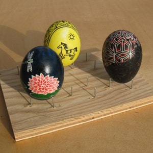 May include: Three decorated Easter eggs on a wooden stand with nails. The eggs are painted with different designs, including a dragonfly, a flower, and a geometric pattern. The stand is made of light wood and has a natural finish.