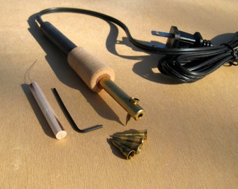 May include: An electric wood burning tool with a black and brown cord and plug. The tool has a wooden handle and a metal tip. The tool comes with a metal rod, a small allen wrench, and five metal tips.