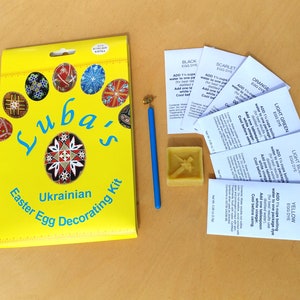 Puede incluir: Una caja amarilla con el texto "Luba's Ukrainian Easter Egg Decorating Kit" en ella. La caja está rodeada de varios paquetes de tinte para huevos de colores, un crayón de cera azul y un bloque de cera.