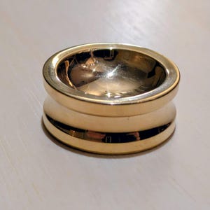May include: A gold-colored, cylindrical object with a rounded top and a wider base. The object has a smooth, reflective surface, and appears to be made of metal. The design features a concave top and a central, rounded section.