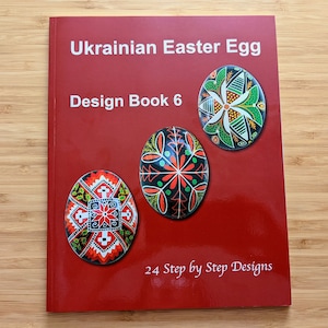 Pode incluir: Um livro vermelho intitulado "Ukrainian Easter Egg Design Book 6" com imagens de ovos decorados. A capa apresenta três ovos pysanky coloridos e o texto "24 Step by Step Designs". O livro está sobre uma superfície de madeira castanha clara.