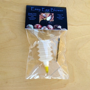 Peut inclure: Un souffleur à œufs en plastique blanc avec une pointe jaune et une aiguille, emballé dans un sac en plastique transparent avec une étiquette noire et blanche qui indique "Easy Egg Blower". L'étiquette comprend également des instructions d'utilisation et de nettoyage.