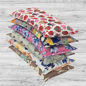 May include: A stack of seven rectangular fabric pouches featuring various patterns including chocolate-covered strawberries, blue floral prints, cartoon characters, and botanical designs in shades of pink, blue, yellow, green, and brown.