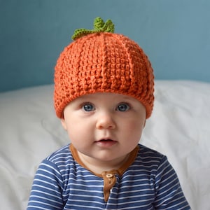 May include: A bright orange crochet beanie hat designed to resemble a pumpkin, featuring vertical ribbed texture and a small green crocheted stem and leaf detail at the top center. The hat is crafted with a thick yarn in a pumpkin-like shape.