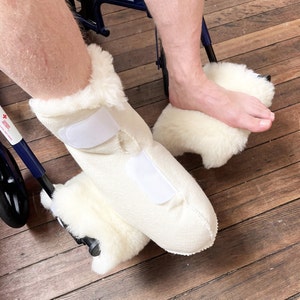 Pode incluir: Um par de protetores de botas brancos e felpudos, que chegam ao tornozelo, com fechos de velcro. Os protetores de botas são usados por uma pessoa sentada numa cadeira de rodas.