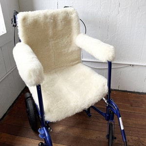 Peut inclure: Une housse de fauteuil roulant en peau de mouton blanche avec un cadre et des roues en métal bleu. La housse comprend des accoudoirs et un coussin d'assise.