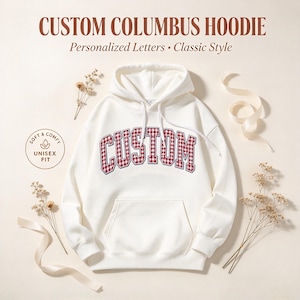 Peut inclure: Un sweat à capuche blanc avec le mot CUSTOM écrit en lettres bloc à carreaux vichy rouges et blancs sur la poitrine. Le sweat comprend une poche kangourou, des manches longues et des cordons blancs. Texte : CUSTOM COLUMBUS HOODIE.
