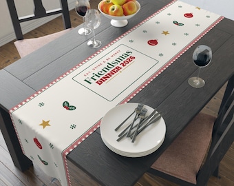 Puede incluir: Un camino de mesa color crema con un borde a cuadros rojos y blancos y un patrón repetitivo de adornos rojos, estrellas amarillas y acebo verde. El texto central dice: EAT, DRINK & BE MERRY FRIENDS-MAS DINNER 2026.