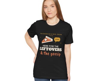 May include: A black short-sleeve t-shirt featuring the text FRIENDSGIVING CREW 2024, a slice of pie, a speech bubble, and the text HERE FOR THE LEFTOVERS & the gossip in white and orange lettering on the front center of the garment.