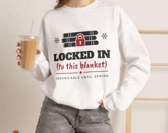 May include: A white long-sleeve sweatshirt featuring a graphic of a red padlock over black bars, with the text "LOCKED IN (to this blanket)" and "UNAVAILABLE UNTIL SPRING" in black and red, accented by small snowflake icons.