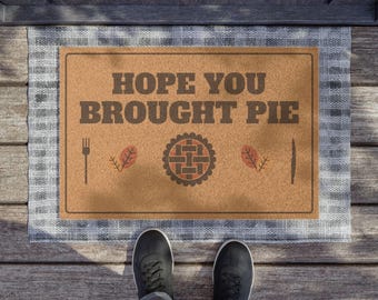 Puede incluir: Un felpudo rectangular de color tostado con un borde de cuadros grises que presenta el texto HOPE YOU BROUGHT PIE en letras marrón oscuro. El diseño incluye un gráfico central de una tarta flanqueado por un tenedor a la izquierda y un cuchillo a la derecha.
