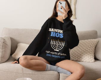 Puede incluir: Una sudadera negra de manga larga con el texto "HANUKKAH HOST" en blanco y azul, un gráfico de una menorá blanca con llamas doradas y el texto "eight nights of latkes, ZERO NIGHTS OFF" en letras blancas y azules.