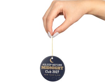 Puede incluir: Un adorno redondo de cerámica azul oscuro con una luna creciente dorada y el texto ASLEEP BEFORE MIDNIGHT CLUB 2027 NEW YEAR'S EVE en letras doradas y blancas, suspendido por un fino cordón dorado sobre un fondo blanco liso.