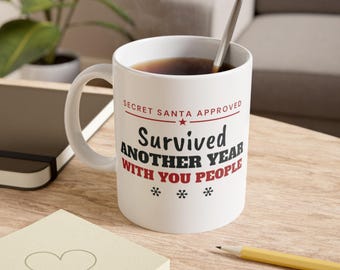 May include: A white ceramic mug with a handle, filled with dark liquid and a silver spoon. The text reads: SECRET SANTA APPROVED, Survived ANOTHER YEAR WITH YOU PEOPLE, with three small snowflake icons printed in black and red ink.