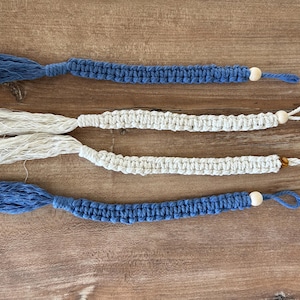 May include: Four macrame bookmarks arranged horizontally. Two are blue and two are off-white. Each bookmark features a knotted cord design, a tassel at one end, a loop at the other, and a single round wooden bead near the loop for decoration.