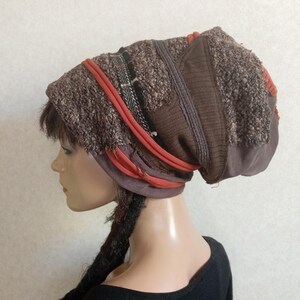 May include: A slouchy beanie hat in shades of brown, featuring a textured, nubby knit fabric combined with smooth brown panels and accented by horizontal bands of burnt orange fabric and a thin, dark patterned trim.