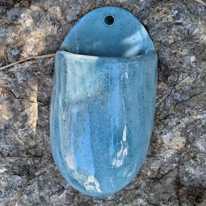 May include: A wall-mounted ceramic planter with a glossy light blue glaze and dark blue speckles. The vessel features a rounded, organic shape with vertical indentations along the body and a single circular hole at the top for hanging.