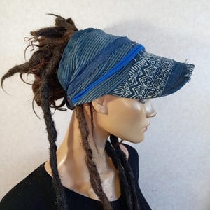 Può includere: Una visiera blu con corona in velluto a coste testurizzato e visiera con motivo geometrico bianco. Il cappello presenta un dettaglio con fascia elastica blu brillante e bordi sfrangiati, indossato sopra lunghi dreadlock castani.