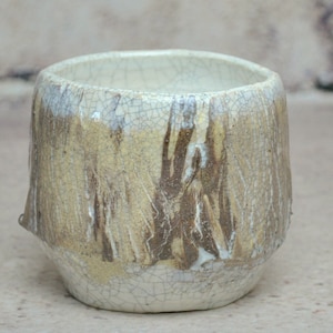 Peut inclure: Une petite tasse en céramique blanche avec un glaçage craquelé et des accents brun et or. La tasse a une forme arrondie et un bord légèrement évasé.
