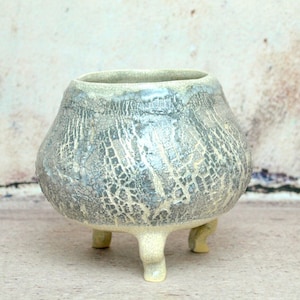 Peut inclure: Un pot en céramique avec un glaçage craquelé gris clair et blanc. Le pot a trois pieds et un corps arrondi.