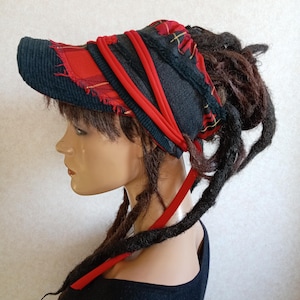 Può includere: Una visiera con tesa in velluto a coste nero e corona in tessuto scozzese rosso e nero. Il design presenta fasce di tessuto rosso sovrapposte e bordi sfrangiati, con lunghi nastri rossi che pendono dalla parte posteriore del copricapo.