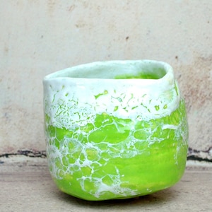Puede incluir: Una taza de cerámica verde con esmalte craquelado blanco. La taza tiene una forma redondeada y un borde ancho.