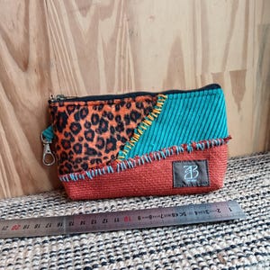 Peut inclure: Petite pochette rectangulaire zippée avec un design patchwork. Elle présente des sections d'imprimé léopard, de velours côtelé turquoise et de tissu couleur rouille, cousues avec du fil orange et bleu. Fermeture éclair argentée et petite étiquette.