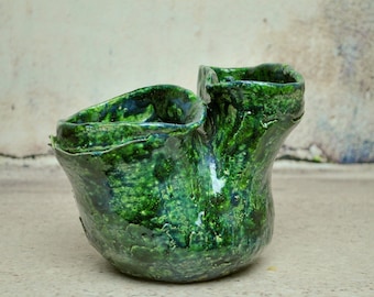 May include: A green ceramic vase with a unique, organic shape. The vase has two openings, one on each side, and a textured, mottled surface.