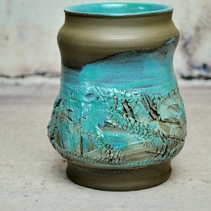 Könnte beinhalten: Eine Keramikvase mit teal-farbener Glasur und einer strukturierten Oberfläche. Die Vase hat einen breiten Boden und einen schmalen Hals.