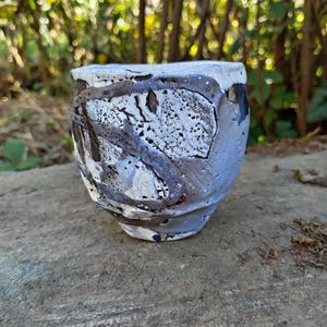 May include: A ceramic cup with a textured, uneven surface featuring a crackled white glaze contrasted by dark, rough, charcoal-coloured patches. The vessel has a rounded, organic shape with a thick rim and a mottled, multi-tonal finish.