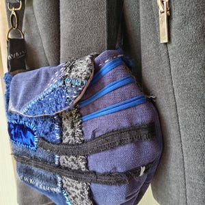 Op de afbeelding: Een patchwork crossbody tas in blauw, grijs en zwart. De tas heeft een klepsluiting en een lange, verstelbare riem. Het ontwerp bevat verschillende texturen en stoffen, waaronder fluweel en gestikte details.