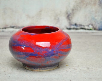 May include: A round ceramic bowl with a vibrant red glaze, accented with streaks of blue. The bowl has a wide opening and a slightly raised base. The glossy finish reflects light, highlighting the colors and textures of the pottery.