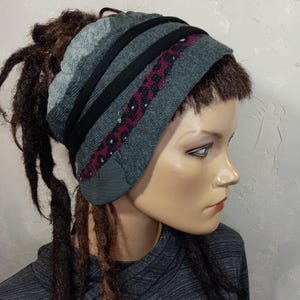 Könnte beinhalten: Ein breites Stirnband mit einem grauen Strickabschnitt, zwei durchgehenden schwarzen horizontalen Bändern und einem dunkelroten Streifen mit schwarzem Leopardenmuster. Das Accessoire wird auf einem Schaufensterpuppenkopf mit braunen Dreadlocks getragen.