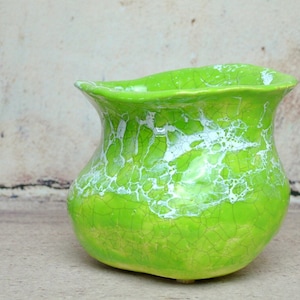 Può includere: Un vaso di ceramica verde con una glassa bianca craquelé. Il vaso ha una base larga e arrotondata e un bordo svasato.