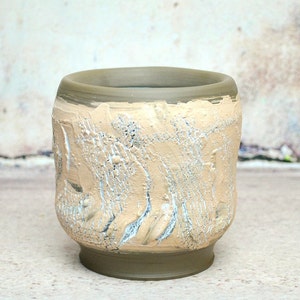 Peut inclure: Un pot en céramique beige avec un glaçage texturé et craquelé. Le pot a un fond arrondi et un haut large et ouvert.