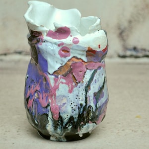 Peut inclure: Un vase en céramique avec un bord irrégulier et ondulé ainsi qu'un émail texturé multicolore. La surface présente un motif de coulures superposées dans des tons de blanc, violet vif, rose soutenu et noir, créant un fini complexe et brillant.