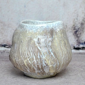 Peut inclure: Une tasse en céramique blanche avec un glaçage craquelé et une surface texturée. La tasse a une forme arrondie et une large ouverture.