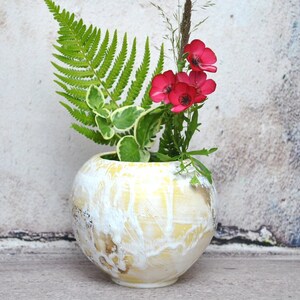 Peut inclure: Un vase en céramique jaune et blanc de forme ronde avec une surface texturée. Le vase est rempli de fleurs roses et de fougères vertes.