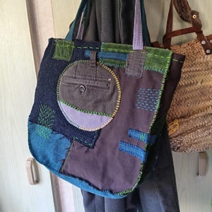 May include: A patchwork tote bag with a variety of blue, green, and brown fabrics. The bag features a circular pocket and decorative stitching in yellow and green. The handles are made of a blue and white checkered fabric. The bag is hanging on a wooden hook.